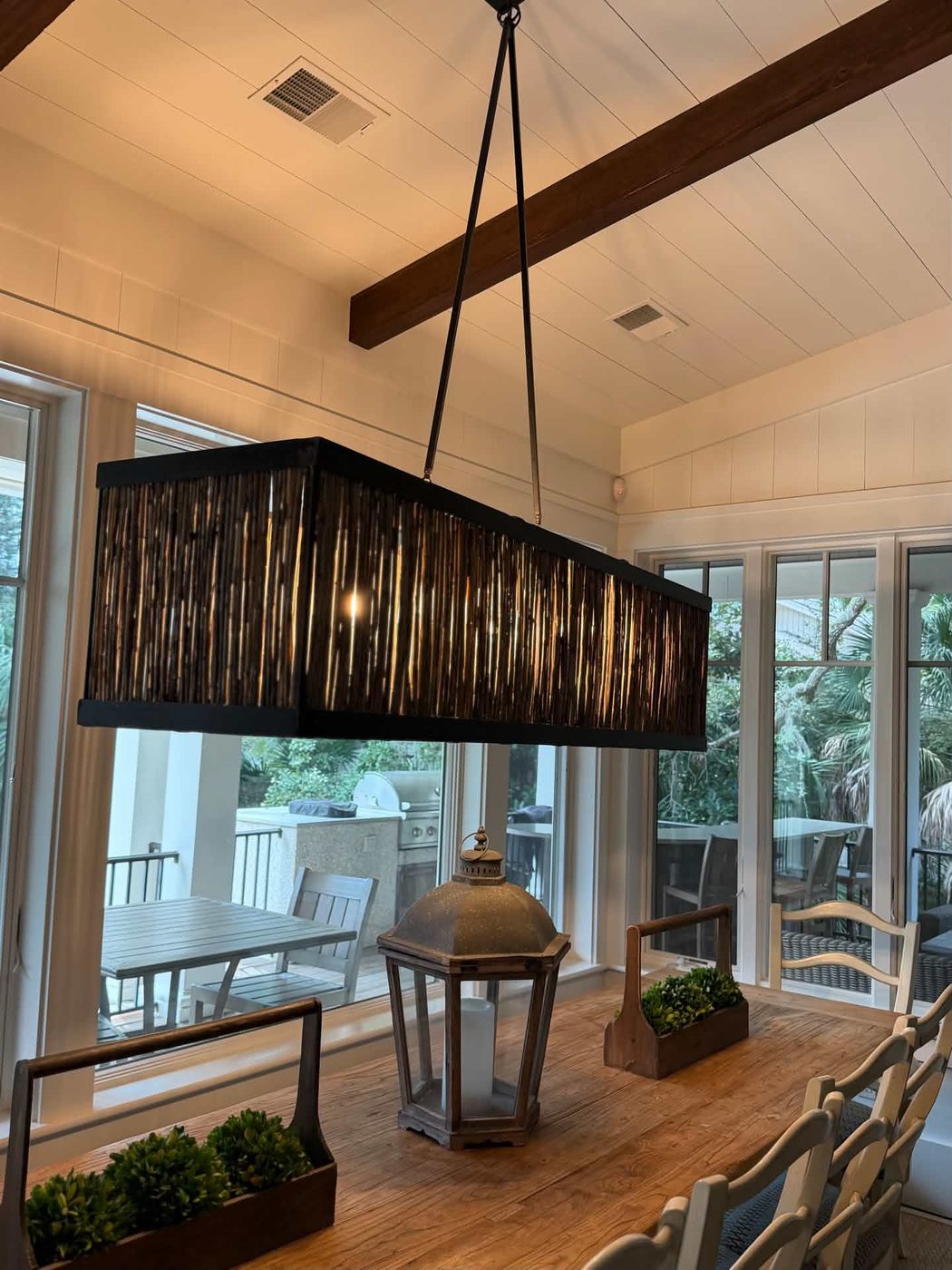 Long rectangular pendant light with a dark reeded shade hung from a ceiling beam above a dining table in a window-lined room