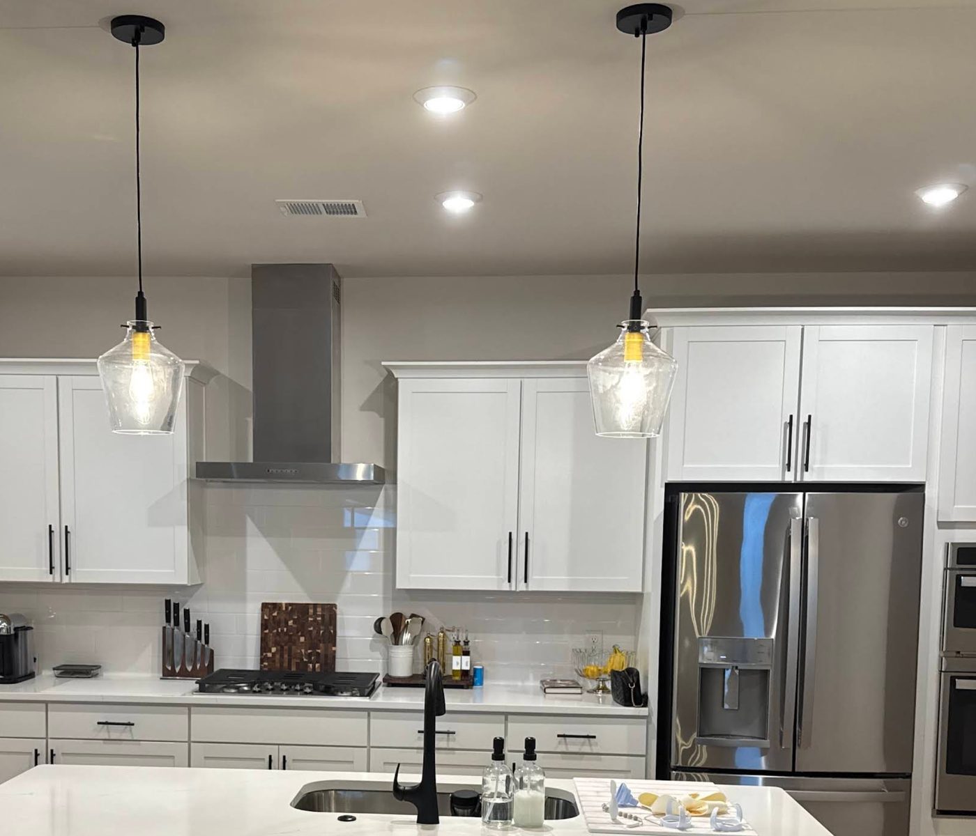 Two glass pendant lights hanging over a kitchen island, with recessed downlights and a stainless steel range hood behind